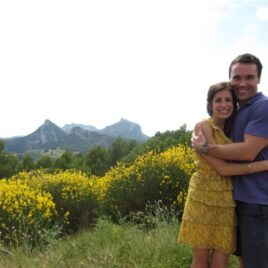 A smiling couple hugs in front of a field of yellow flowers with mountains in the background.