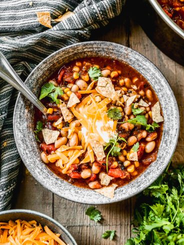 A bowl of taco soup topped with shredded cheese, tortilla chips, cilantro, and sour cream sits on a wooden table.