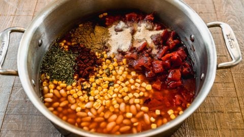 Large pot filled with beans, corn, diced tomatoes, and assorted spices on a wooden surface, ready to be mixed.