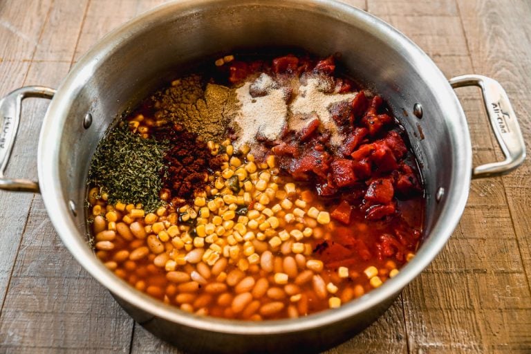 Large pot filled with beans, corn, diced tomatoes, and assorted spices on a wooden surface, ready to be mixed.