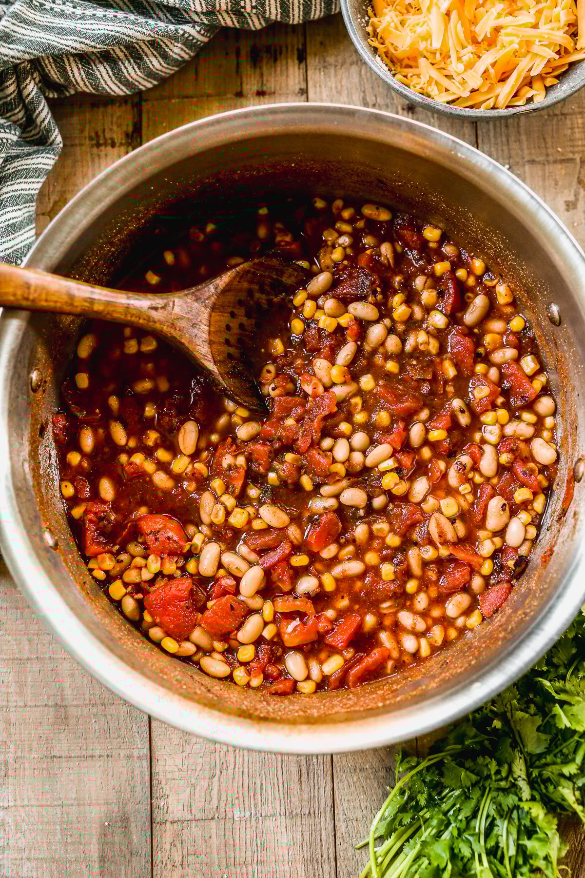 A pot of taco soup with beans, tomatoes, and corn, being stirred with a wooden spoon on a rustic wooden table.