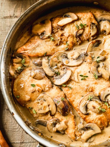 Mushroom chicken in a creamy sauce, garnished with fresh herbs, cooked in a skillet on a wooden surface.