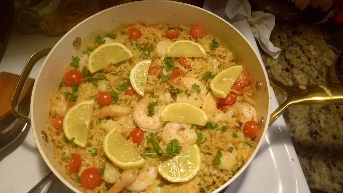 A pan of shrimp and rice with cherry tomatoes, lemon wedges, and chopped herbs on top.