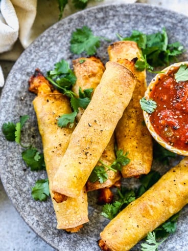 Four crispy chicken taquitos on a gray plate, garnished with cilantro, served with a bowl of salsa.
