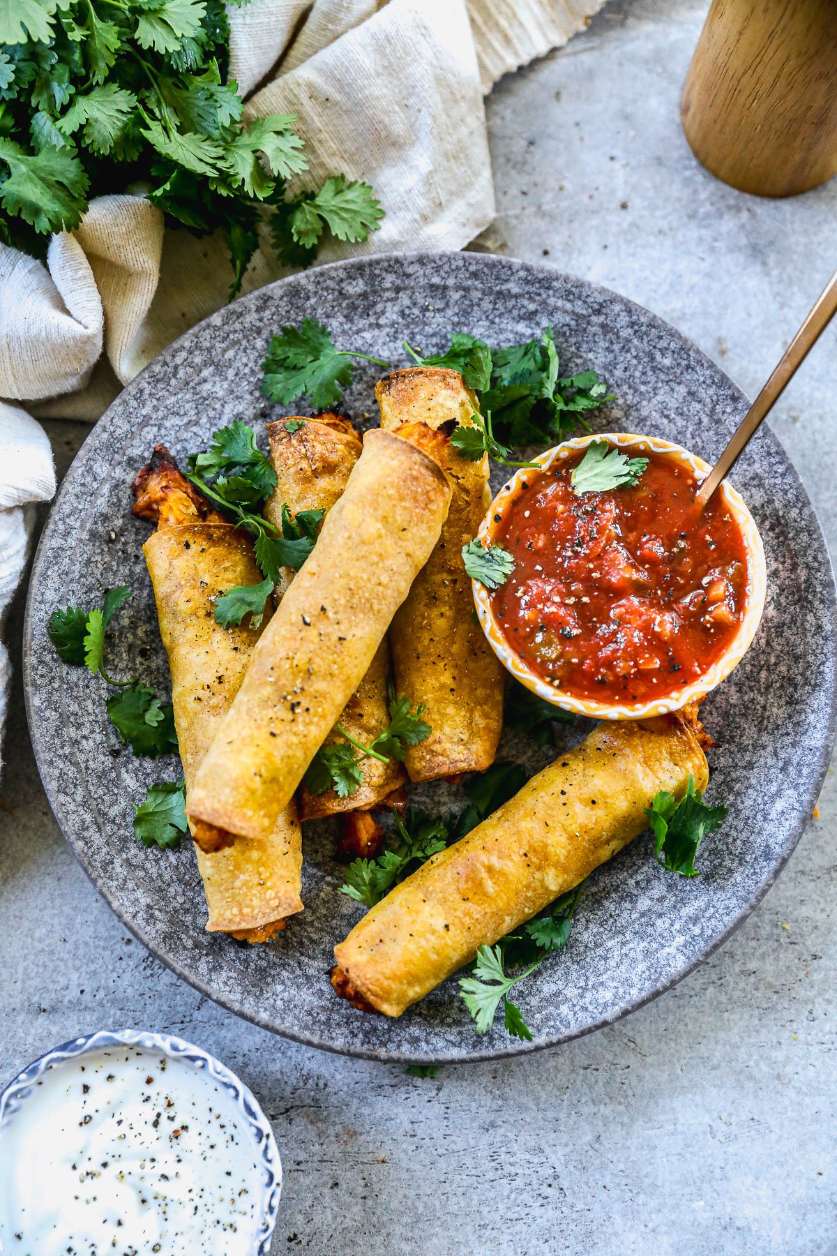A plate of crispy chicken taquitos with fresh cilantro, served with salsa and a small bowl of creamy dip on the side.