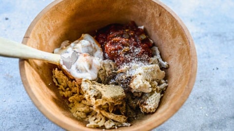 A wooden bowl with shredded chicken sauce, and seasonings being mixed with a wooden spoon.