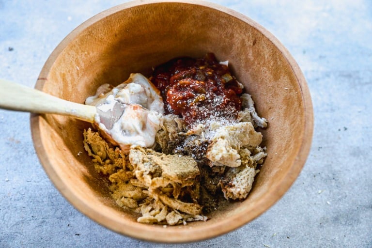 A wooden bowl with shredded chicken sauce, and seasonings being mixed with a wooden spoon.