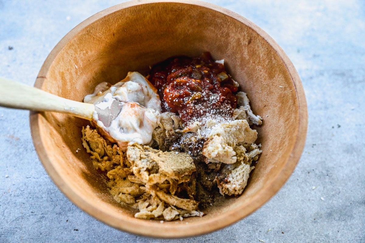 A wooden bowl with shredded chicken sauce, and seasonings being mixed with a wooden spoon.