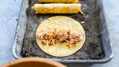 A corn tortilla with shredded chicken filling sits on a baking tray beside two rolled, filled tortillas.