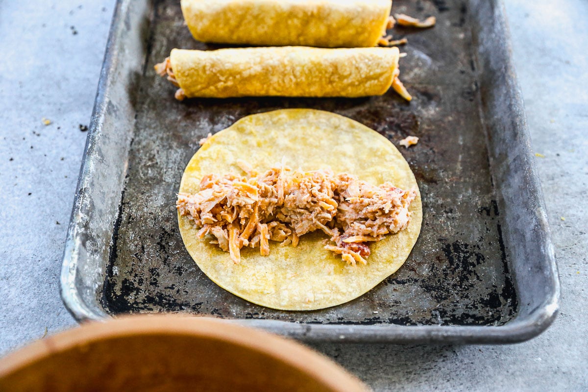 A corn tortilla with shredded chicken filling sits on a baking tray beside two rolled, filled tortillas.