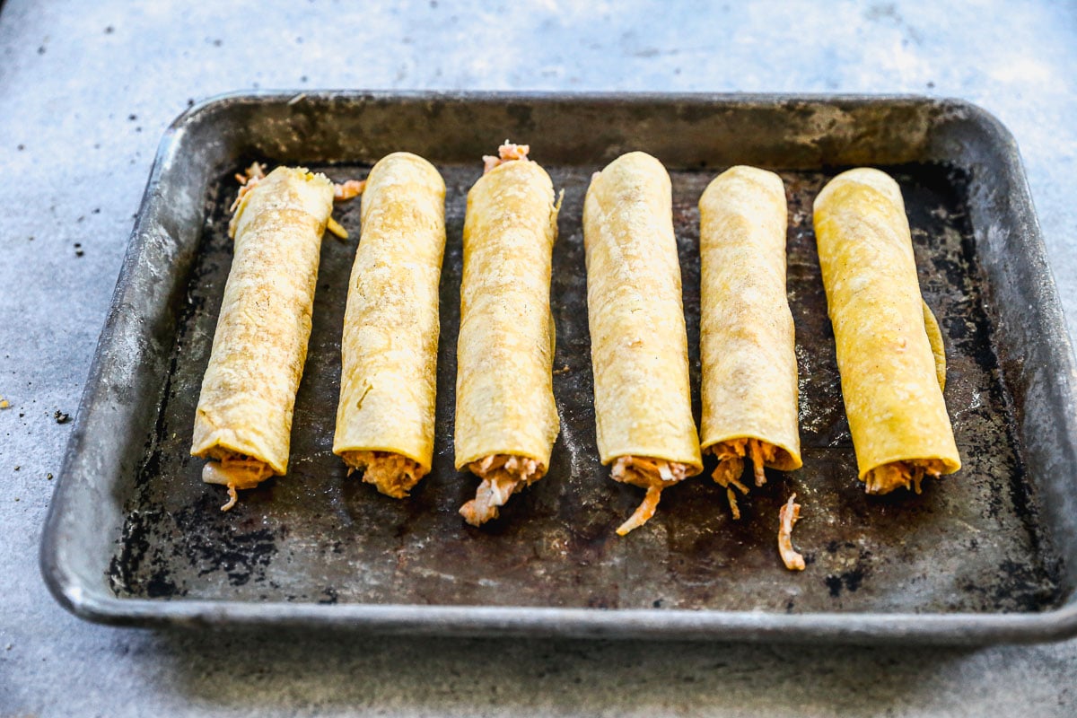 Six rolled corn tortillas filled with shredded chicken on a baking sheet, ready to be cooked.