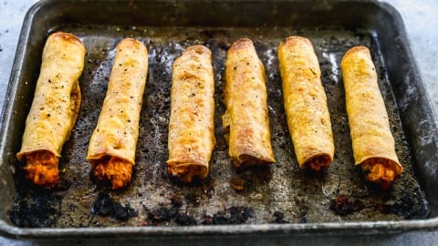 Six baked chicken taquitos filled with orange-colored filling on a worn, dark baking sheet.