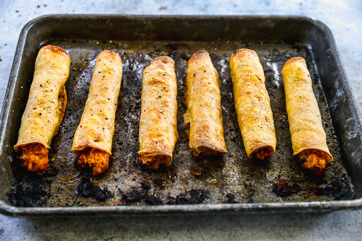 Six baked chicken taquitos filled with orange-colored filling on a worn, dark baking sheet.