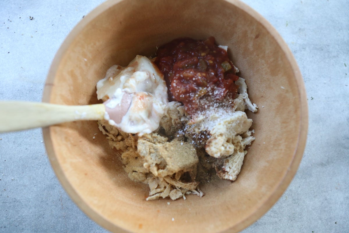 Wooden bowl with shredded chicken, yogurt, salsa, spices, and breadcrumbs, with a wooden spoon on the side.