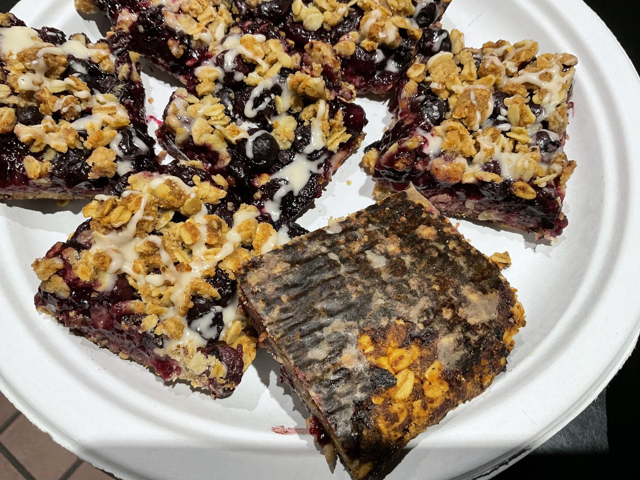 Five berry crumble bars on a plate, one with burnt or overcooked topping, others drizzled with white icing.