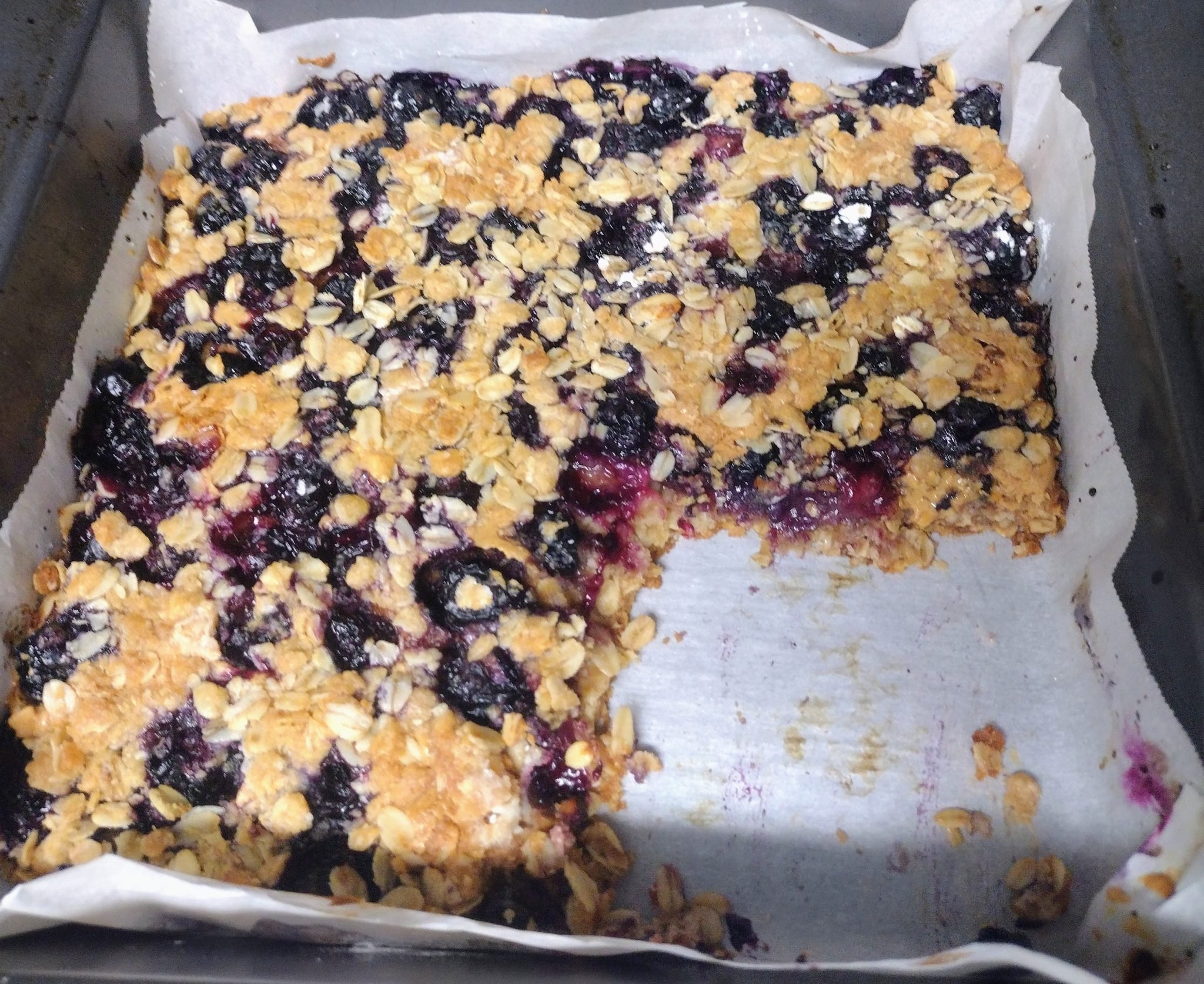 A tray of blueberry oat bars on parchment paper with a square piece missing from the corner.
