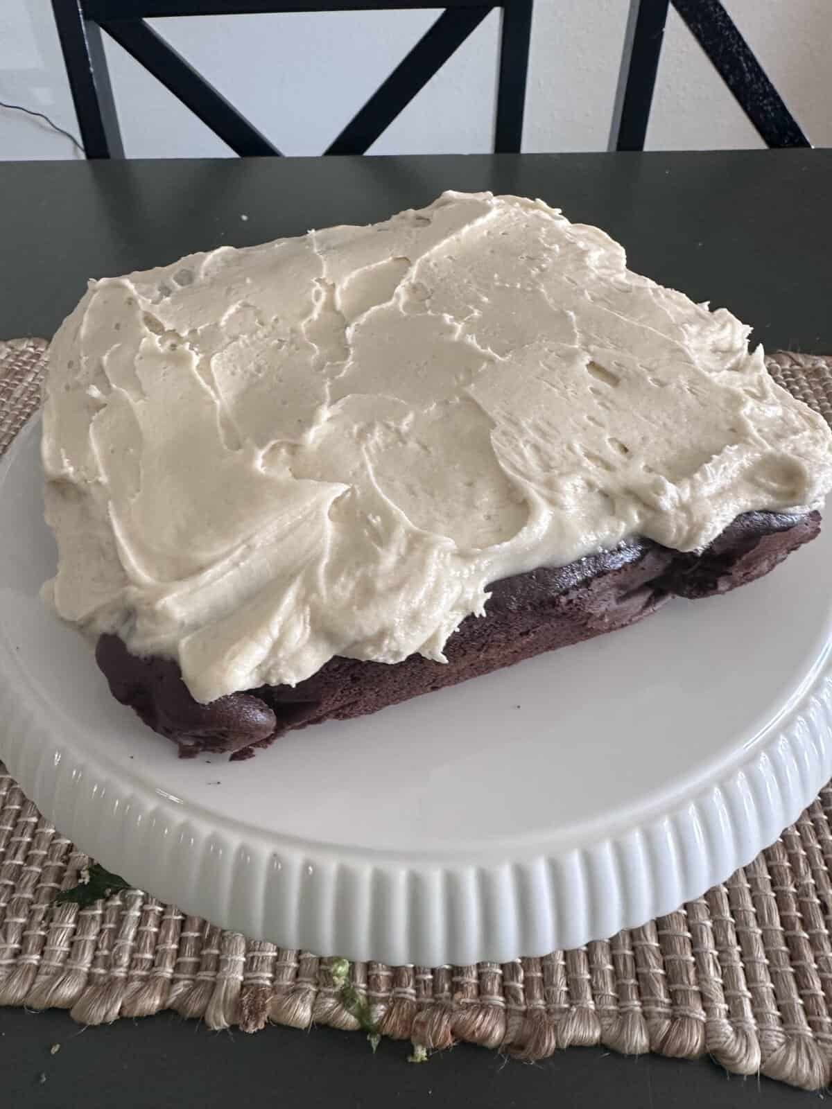 Square chocolate cake with thick white frosting on a white cake stand, on a woven placemat.