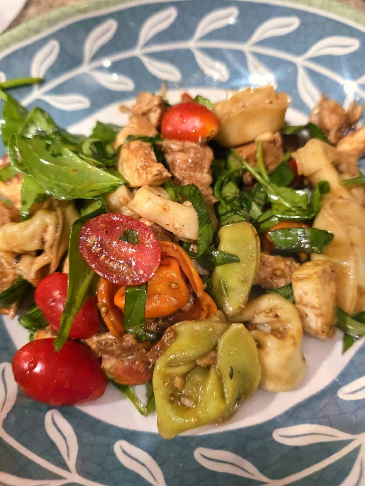 Pasta salad with green tortellini, cherry tomatoes, greens, chicken, and cheese on a patterned plate.