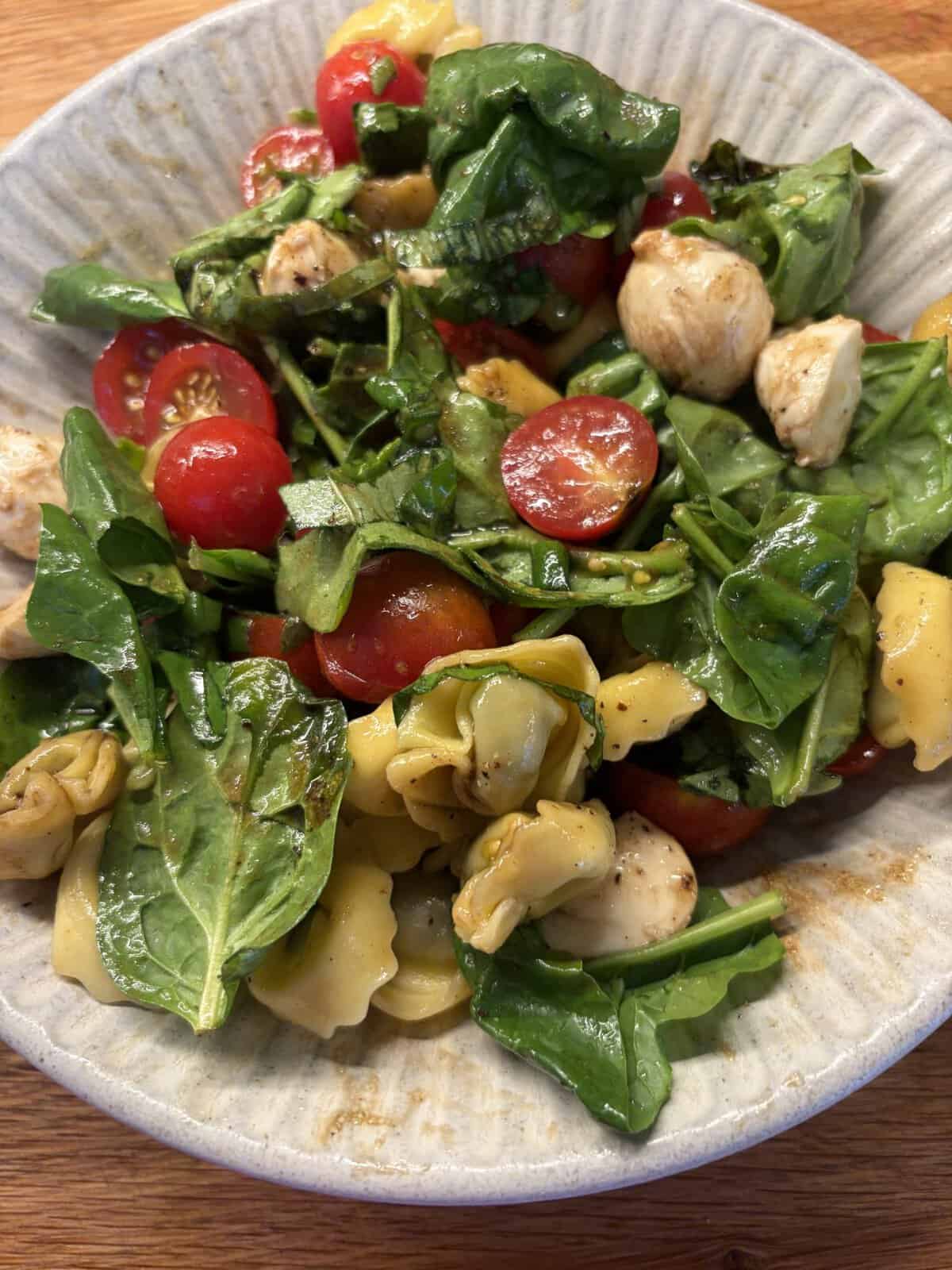 A bowl of tortellini salad with spinach, cherry tomatoes, mozzarella balls, and balsamic dressing.