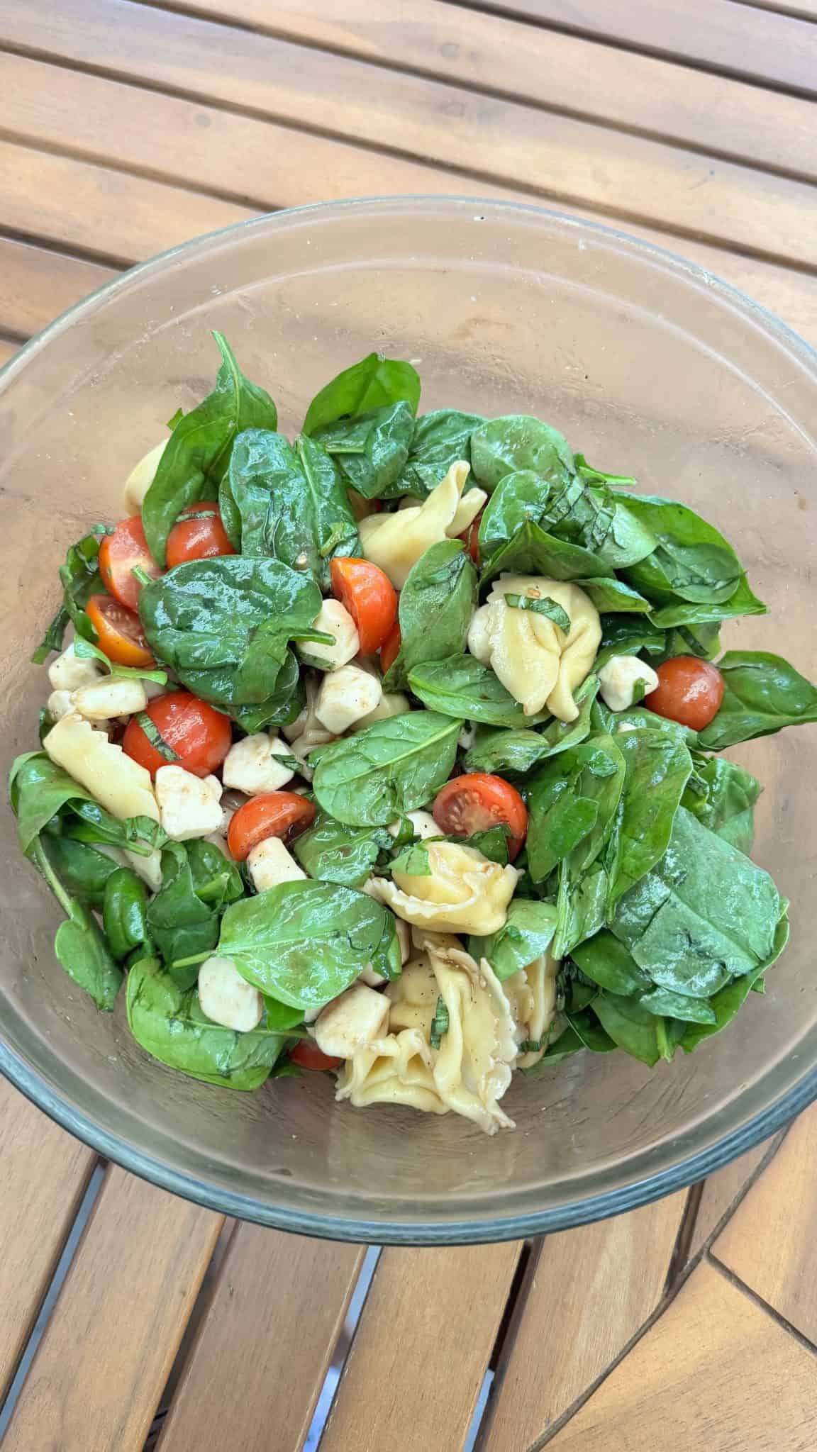 A glass bowl of salad with spinach, cherry tomatoes, mozzarella, and tortellini on a wooden table.