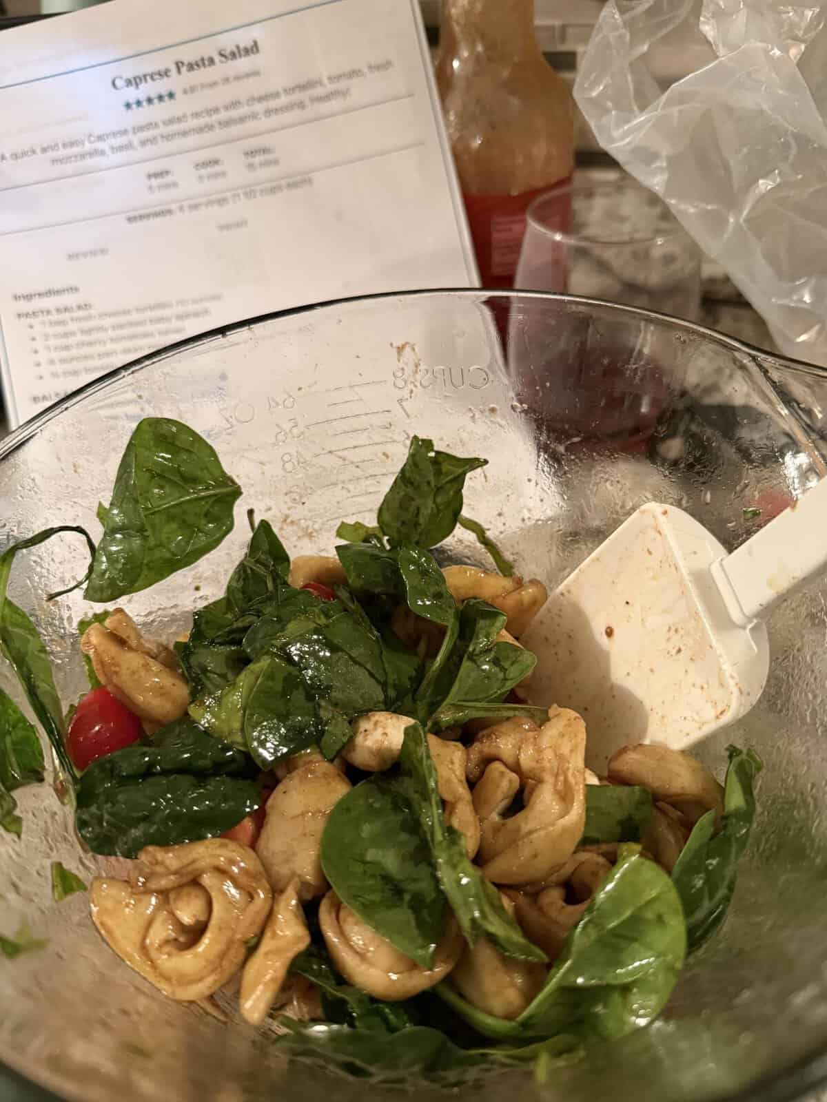 A bowl of caprese pasta salad with spinach, tomatoes, and cheese tortellini, with a recipe and spatula nearby.