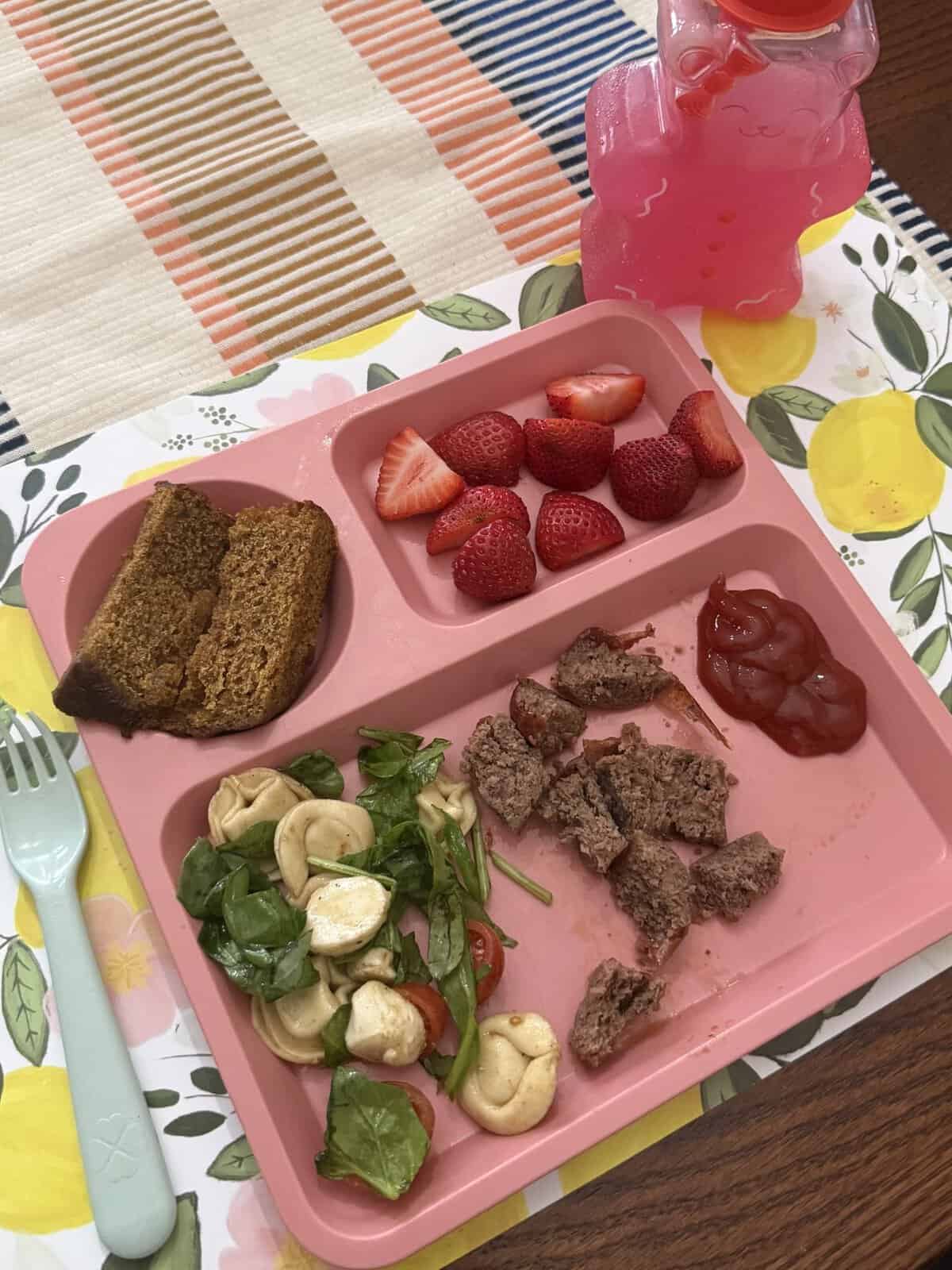 Pink tray with salad, meat pieces with ketchup, strawberries, bread, and a pink drink on a floral placemat.