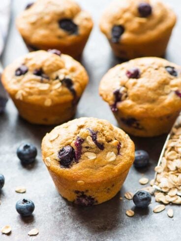 5 healthy blueberry muffins on a granite counter next to a scoop of rolled oats and fresh blueberries