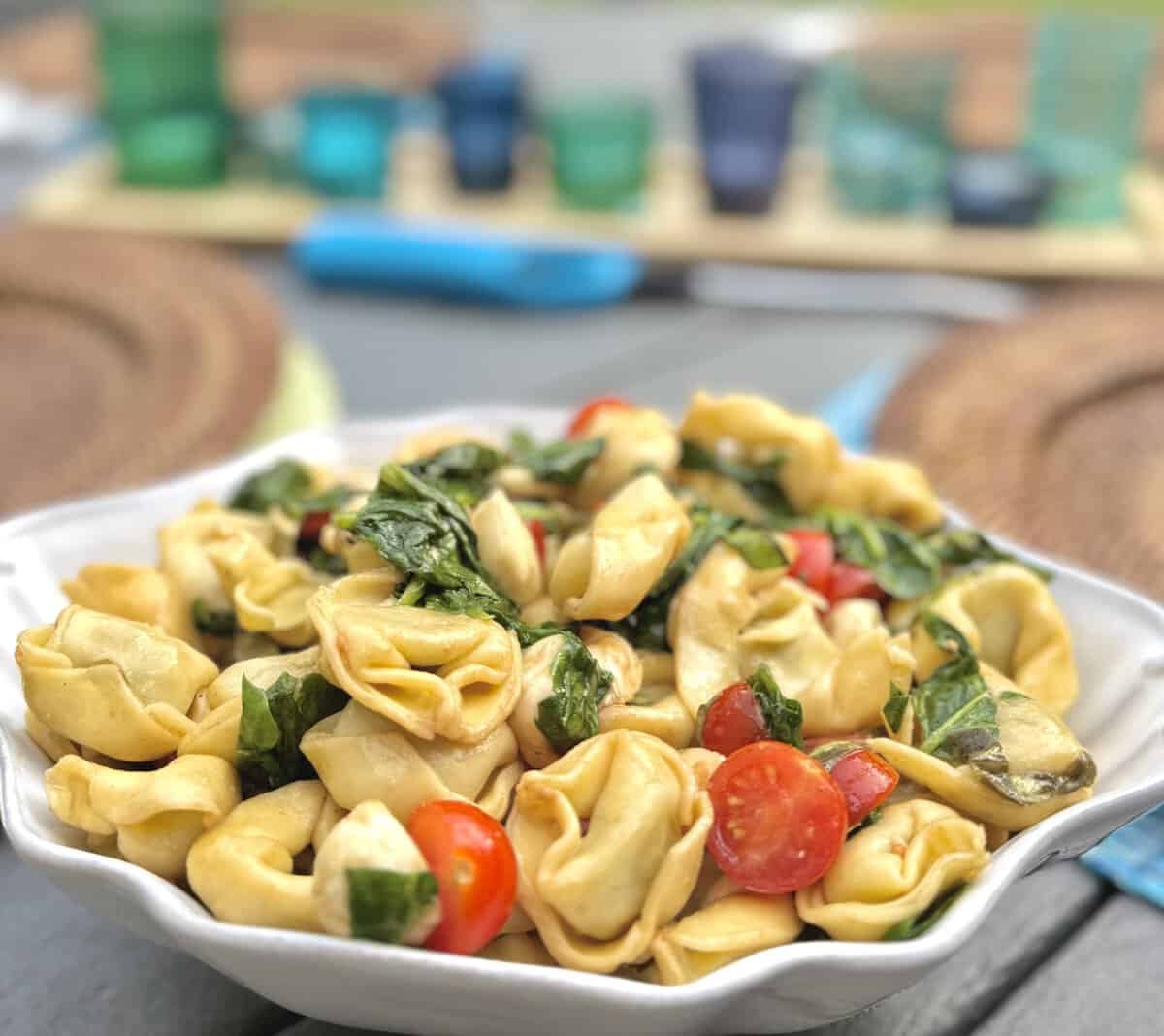 A bowl of tortellini pasta salad with cherry tomatoes and fresh basil sits on an outdoor table.