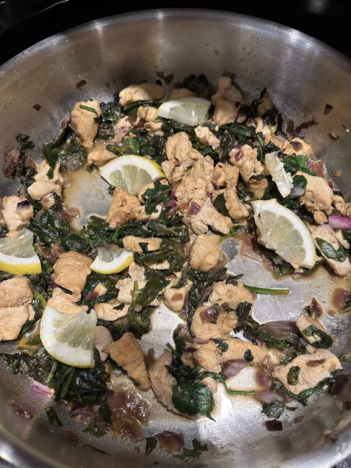 Chicken stir-fry with spinach, red onions, and lemon slices cooking in a stainless steel pan.