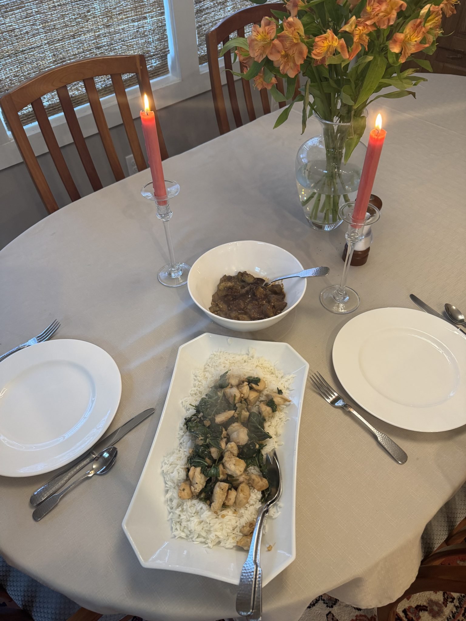 A dinner table with flowers, two lit candles, plates, utensils, rice with chicken and greens, and a bowl of stew.