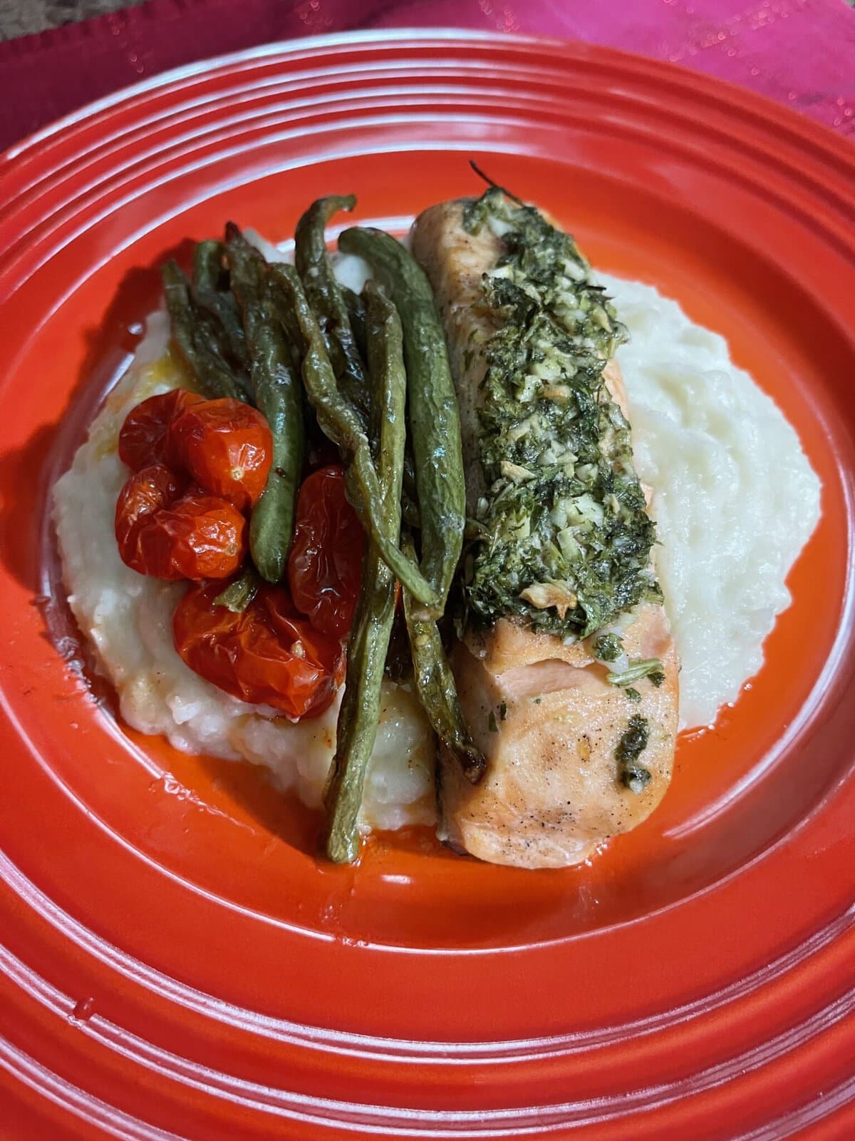 Herb-crusted salmon with green beans, roasted cherry tomatoes, and mashed potatoes on a red plate.