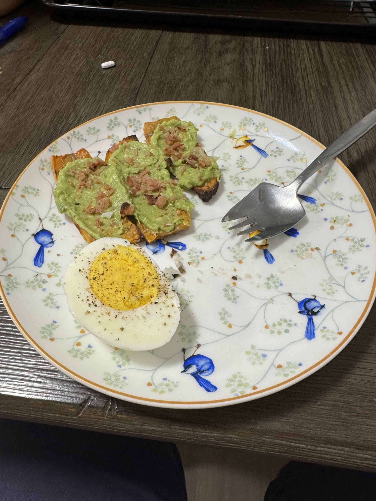 A plate with half a boiled egg and three pieces of toast topped with avocado and tuna, with a fork on the side.