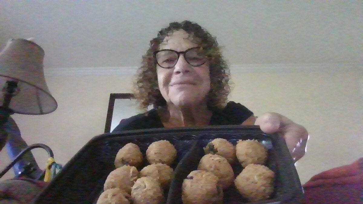 Woman with curly hair and glasses holding a tray of round, fried snacks, smiling at the camera indoors.