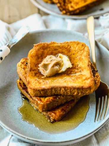 Three slices of pumpkin French toast stacked on a plate with butter and syrup, and a fork and knife beside them.