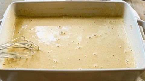 Rectangular dish filled with bubbly, light brown pumpkin custard for French toast; a metal whisk rests on the left side.