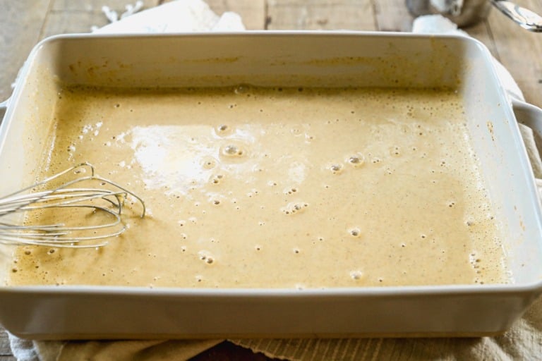 Rectangular dish filled with bubbly, light brown pumpkin custard for French toast; a metal whisk rests on the left side.