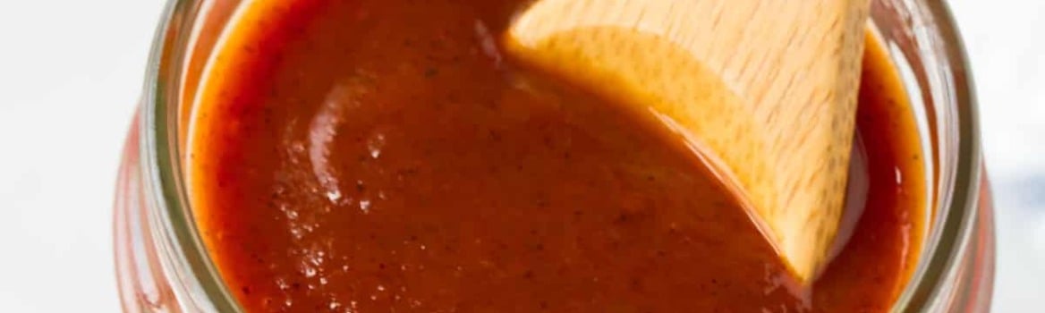 A jar of barbecue sauce with a wooden spoon inside, placed on a white surface with a blue-checkered towel nearby.