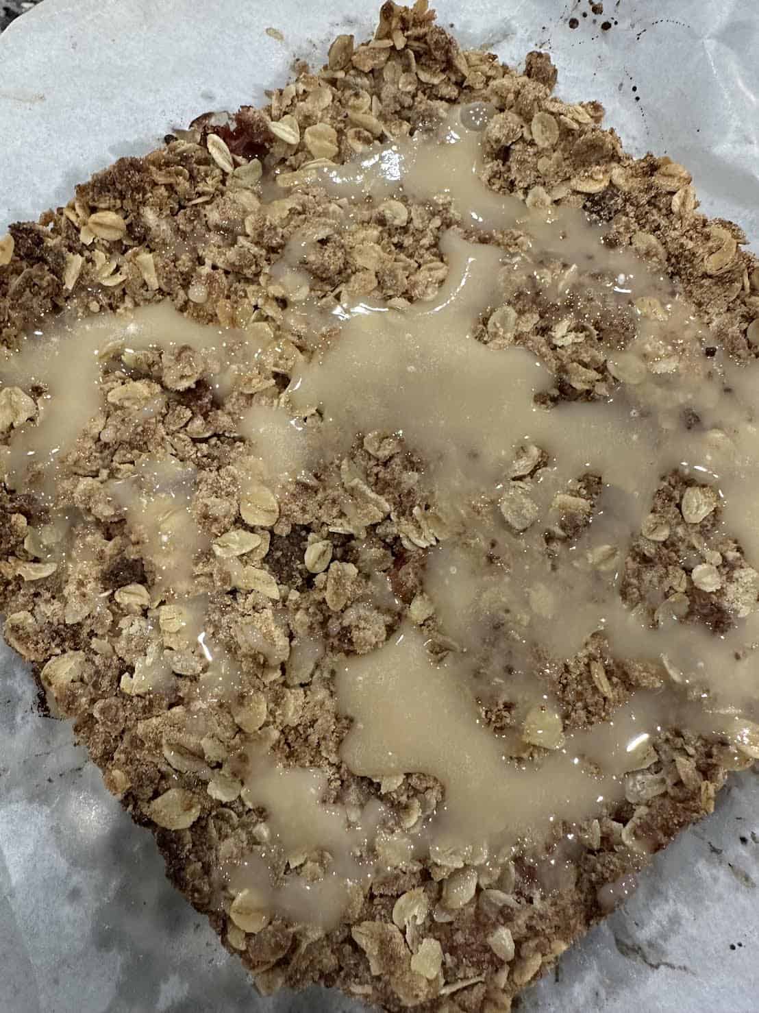 A square oat crumble dessert with glaze drizzled on top, placed on parchment paper.