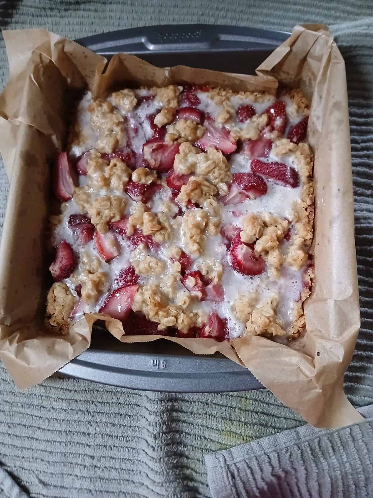A baked strawberry crumble bar sits in a parchment-lined square pan on a striped kitchen towel.
