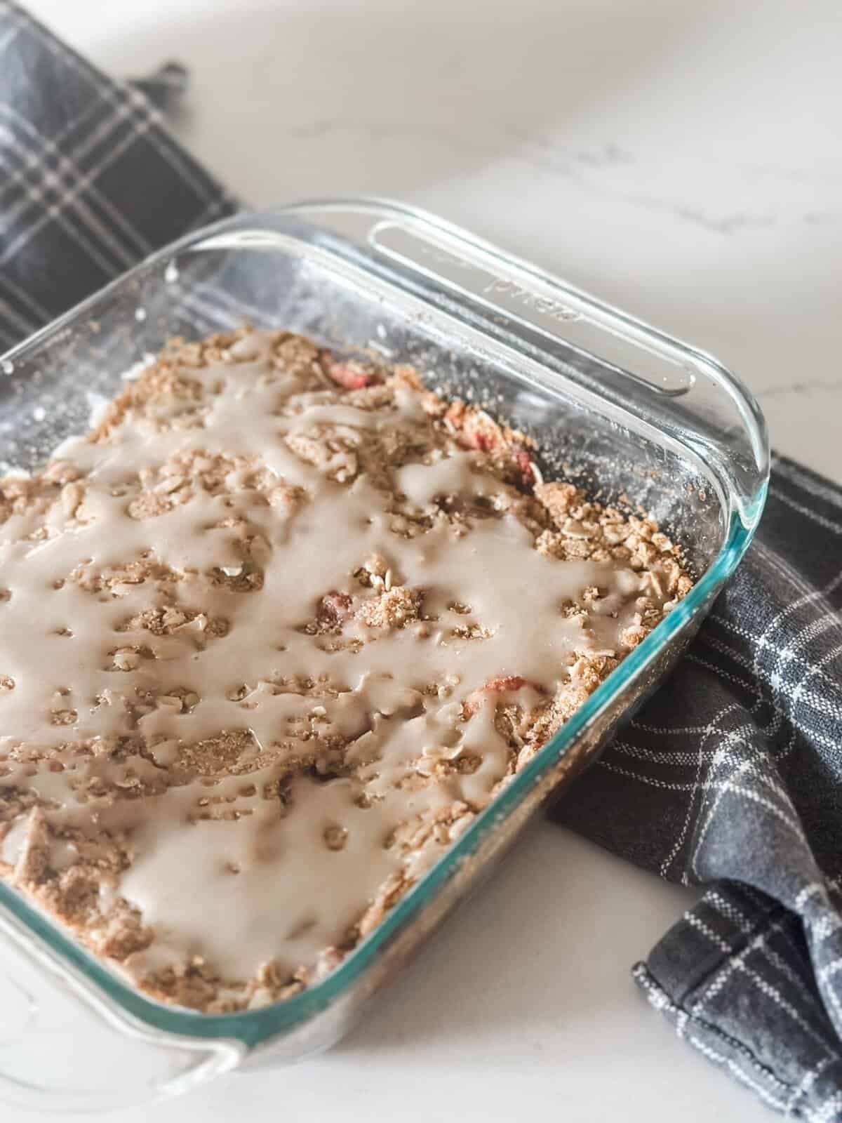 A glass baking dish filled with freshly baked dessert, topped with a light drizzle of glaze, set on a marble countertop. A dark plaid cloth napkin is placed beside it.