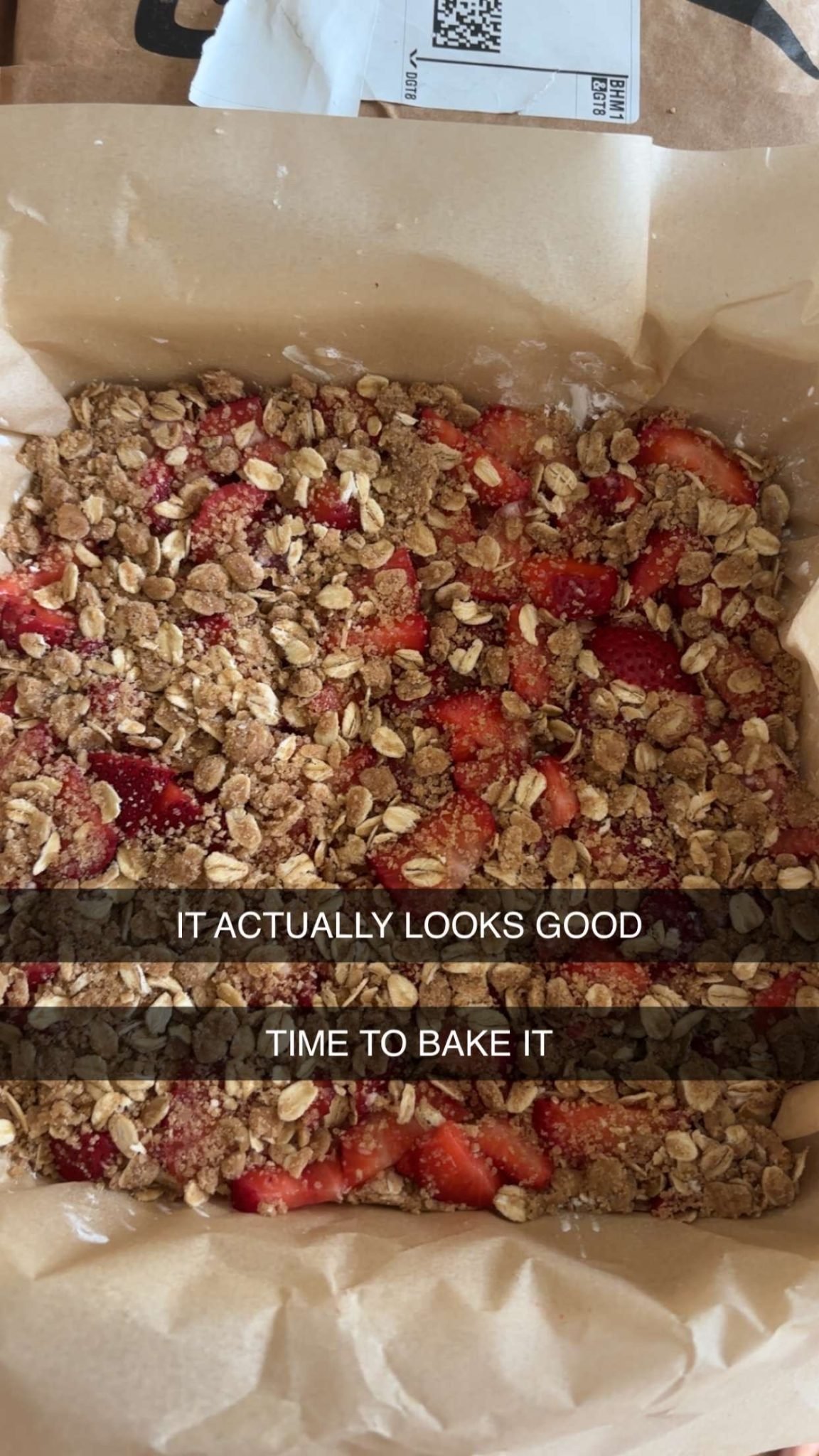 A baking pan lined with parchment paper holds an unbaked strawberry crumble topped with oats and brown sugar. Text overlay reads, "IT ACTUALLY LOOKS GOOD TIME TO BAKE IT.