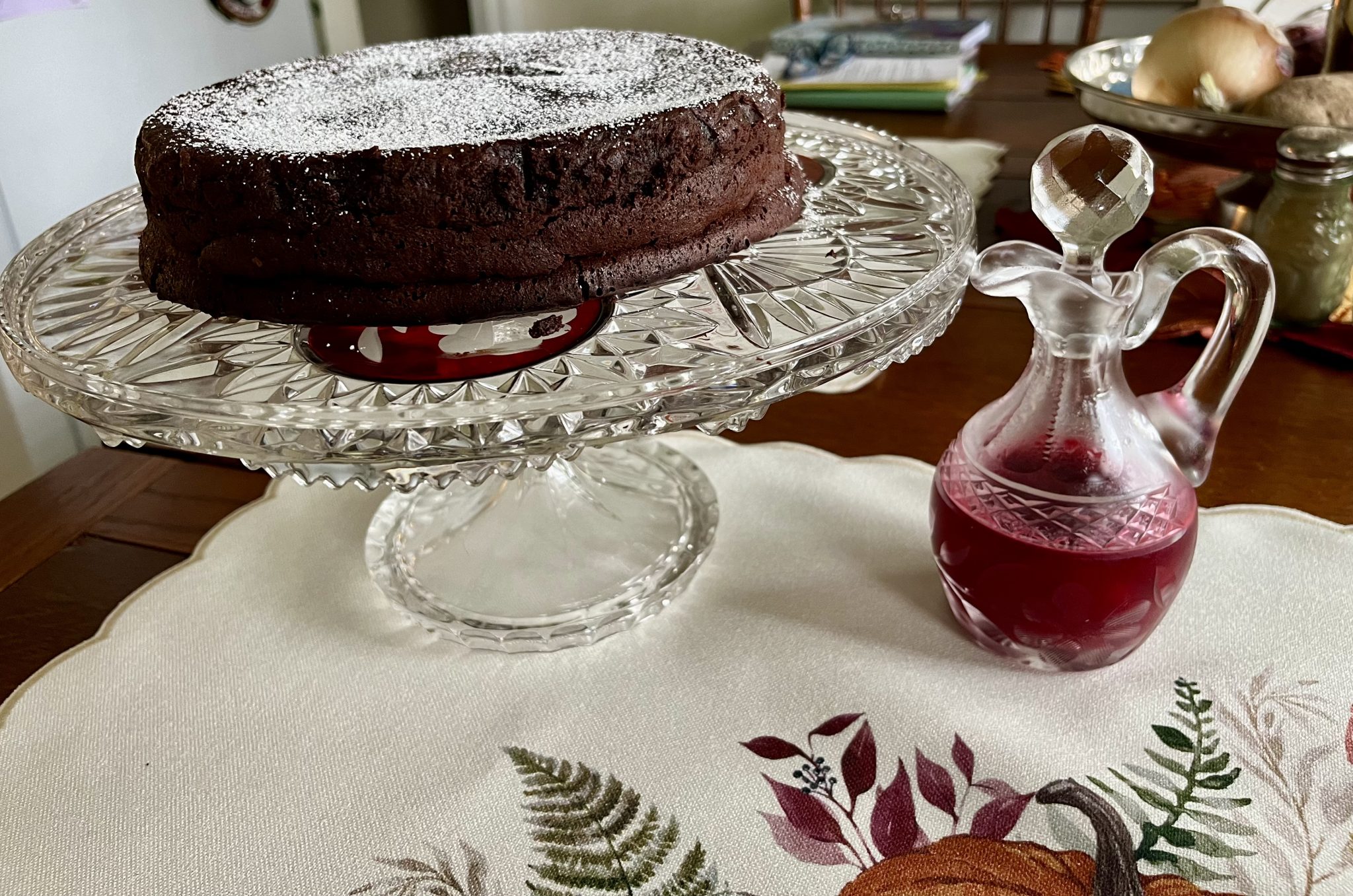 A chocolate cake on a glass stand and a glass carafe of red liquid sit on a table with a decorative placemat.