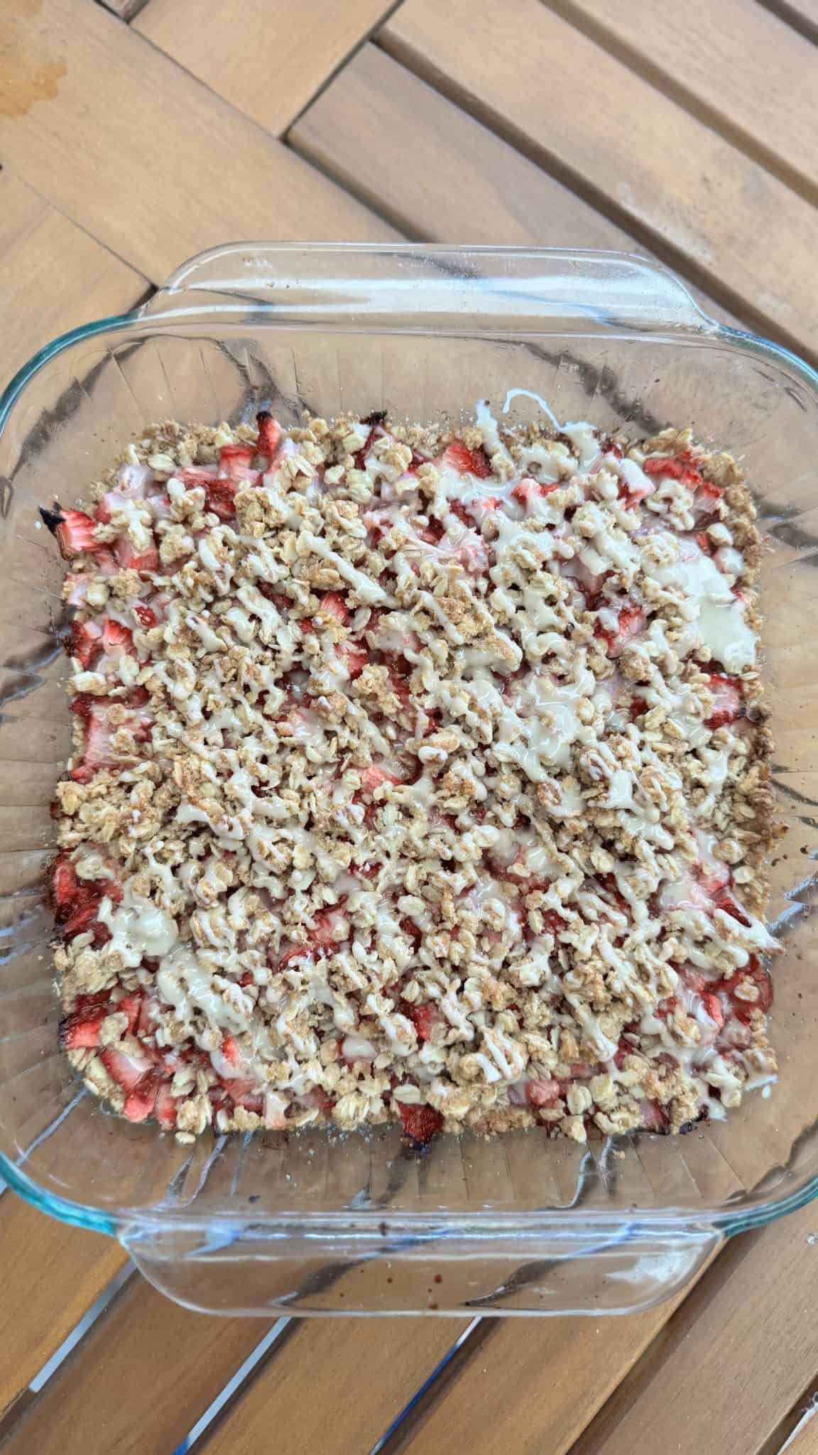 A glass baking dish filled with strawberry crumble topped with a white glaze, on a wooden table.