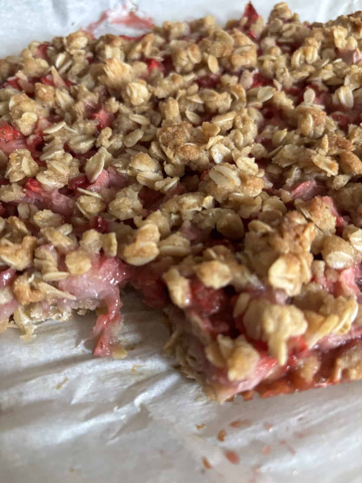 A close-up of a fruit oat crumble bar with a piece missing, showing a gooey fruit filling and crumbly oat topping.