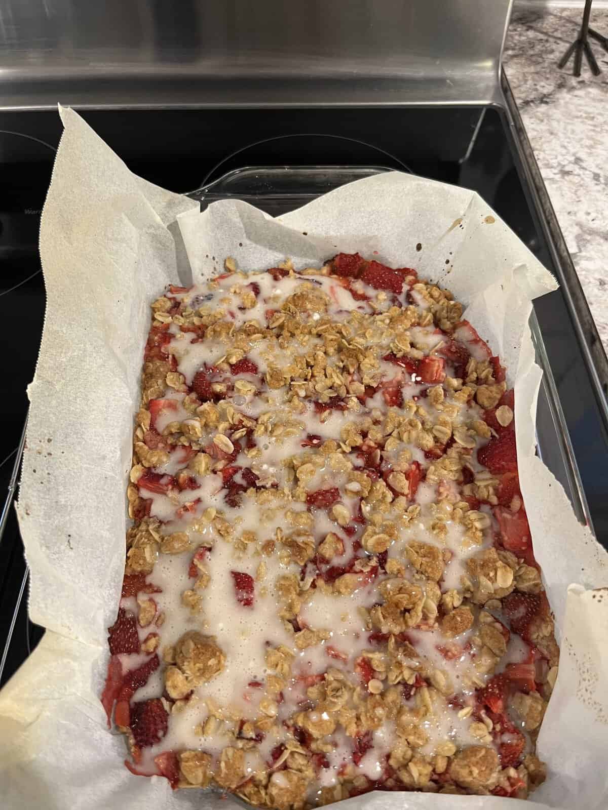 A baked strawberry oat crumble with icing, lined with parchment paper, sits in a glass dish on a kitchen counter.