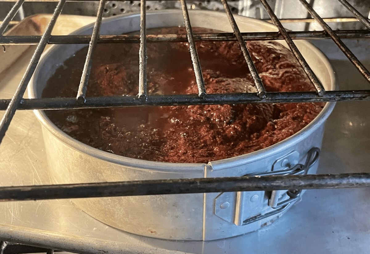 Chocolate cake baking in a round metal pan inside an oven, viewed through the oven rack.