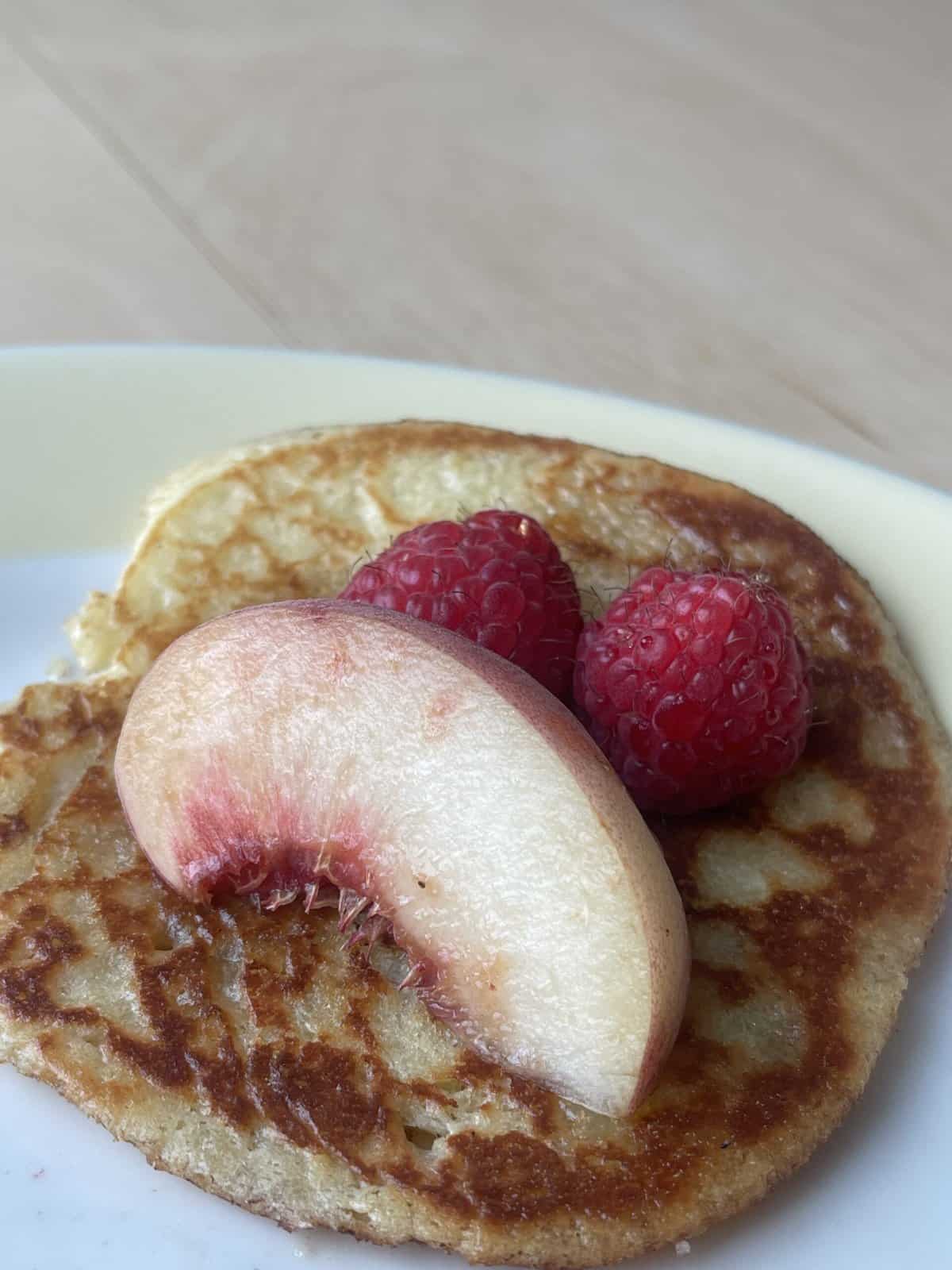 A golden pancake topped with a peach slice and two raspberries on a white plate.