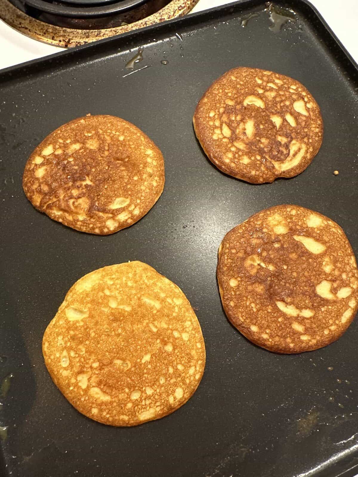 Four golden brown pancakes cooking on a black griddle near a stove burner.