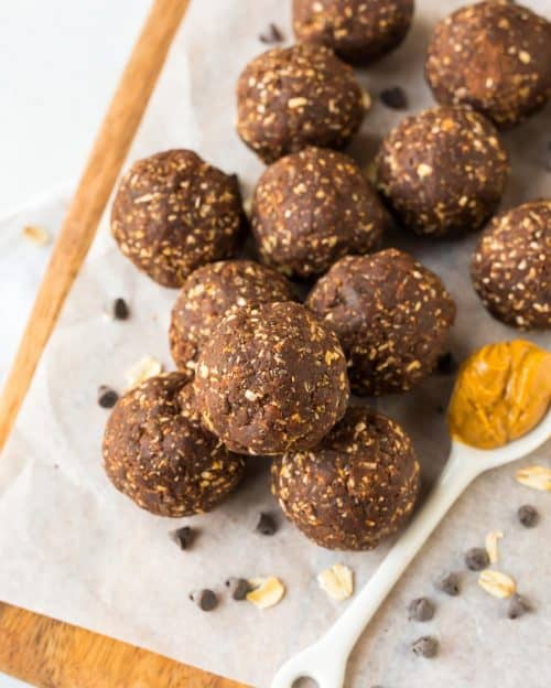 Chocolate oat energy balls stacked on parchment paper, with a spoonful of peanut butter and chocolate chips nearby.