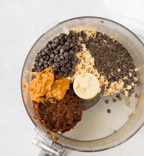 A food processor with oats, chia seeds, chocolate chips, peanut butter, and a date mixture inside.